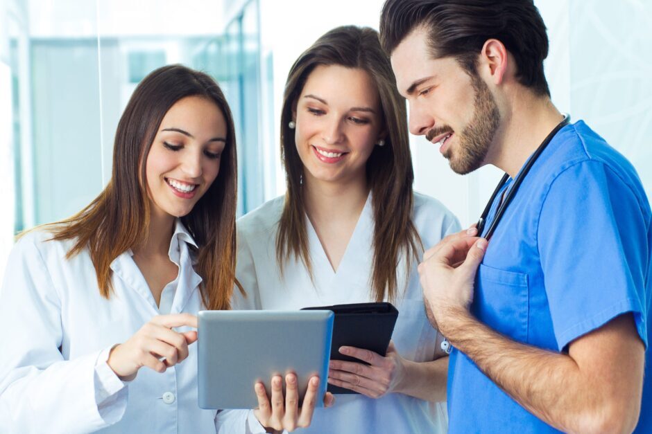 Two female clinical students show their tablets to a male doctor in the hospital corridor.